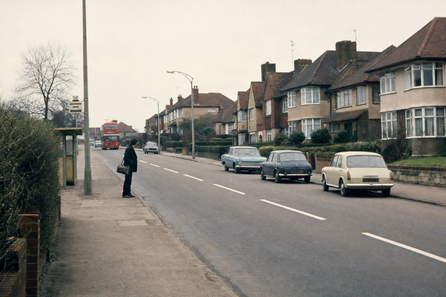 Südenglische Wohnstraße um 1970 mit zeittypischen Häusern, Bushaltestelle, Linksverkehr und einem wartenden jungen Reisenden