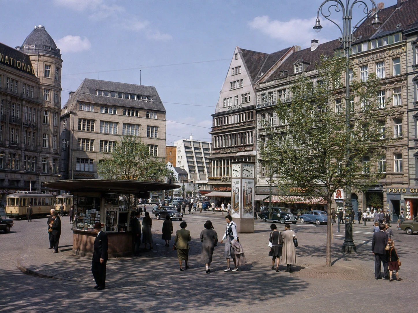 Belebter Kölner Innenstadtplatz mit Straßenbahnen, Kiosk, Geschäften und Passanten in historischer Anmutung